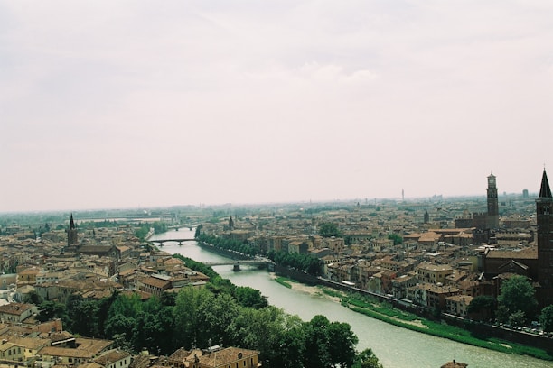 A picturesque urban landscape featuring an expansive view of a historical city with densely packed buildings. A river snakes through the city, bordered by lush greenery. Tall, historical towers and church spires are scattered throughout, standing prominently against the hazy sky.