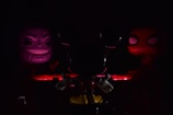 Three character figures are arranged in a dimly lit setting. The central figure is primarily in shadow, characterized by prominent ears and white gloves. On the left, a figure with a sinister grin is illuminated in purple light. To the right, a figure with large, prominent eyes is highlighted in red. The scene is bathed in low light, creating a mysterious and dramatic atmosphere.