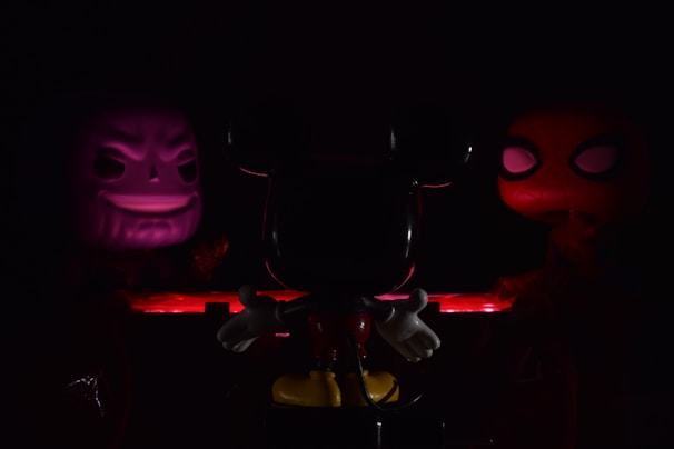 Three character figures are arranged in a dimly lit setting. The central figure is primarily in shadow, characterized by prominent ears and white gloves. On the left, a figure with a sinister grin is illuminated in purple light. To the right, a figure with large, prominent eyes is highlighted in red. The scene is bathed in low light, creating a mysterious and dramatic atmosphere.