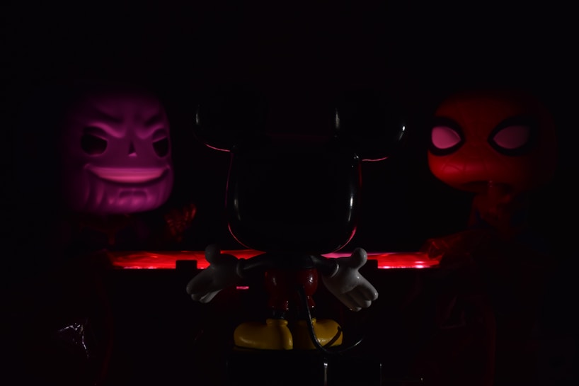 Three character figures are arranged in a dimly lit setting. The central figure is primarily in shadow, characterized by prominent ears and white gloves. On the left, a figure with a sinister grin is illuminated in purple light. To the right, a figure with large, prominent eyes is highlighted in red. The scene is bathed in low light, creating a mysterious and dramatic atmosphere.
