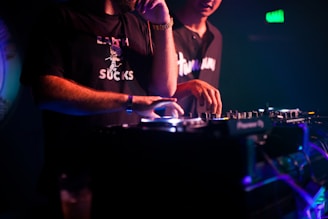 Two individuals are engaged in music mixing, with one adjusting the controls on a mixer in a dimly lit setting. The lighting is primarily purple and blue, creating a vivid nightclub atmosphere. Both are casually dressed, and one of the shirts has text printed on it.