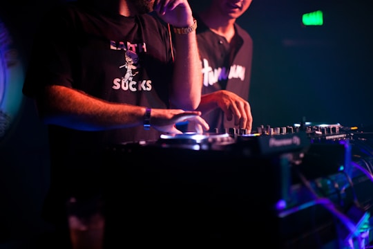 Two individuals are engaged in music mixing, with one adjusting the controls on a mixer in a dimly lit setting. The lighting is primarily purple and blue, creating a vivid nightclub atmosphere. Both are casually dressed, and one of the shirts has text printed on it.