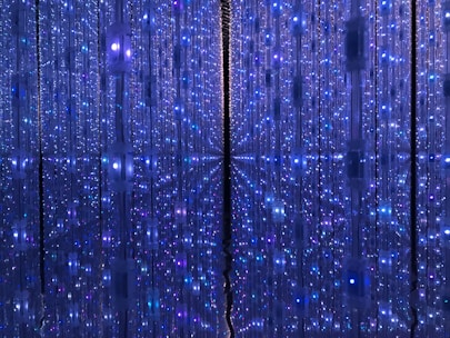 A mesmerizing display of vertical LED light strips creates a tunnel-like effect with reflections that give a sense of infinite depth. The lights are predominantly blue, interspersed with hints of purple and white, evoking a futuristic and otherworldly atmosphere.