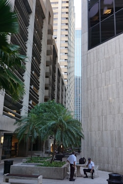 Tall skyscrapers surround a small courtyard area with palm trees. Two uniformed security personnel are conversing in the foreground. The architecture features modern and sleek designs with concrete and glass elements.