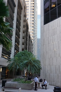 Tall skyscrapers surround a small courtyard area with palm trees. Two uniformed security personnel are conversing in the foreground. The architecture features modern and sleek designs with concrete and glass elements.