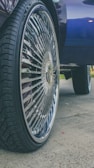 A close-up of a car wheel with a large, shiny rim featuring intricate designs. The tire has a deep tread pattern, and the background includes part of the car body and a hint of grass and pavement.