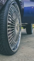 A close-up of a car wheel with a large, shiny rim featuring intricate designs. The tire has a deep tread pattern, and the background includes part of the car body and a hint of grass and pavement.