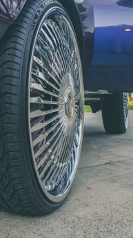 A close-up of a car wheel with a large, shiny rim featuring intricate designs. The tire has a deep tread pattern, and the background includes part of the car body and a hint of grass and pavement.