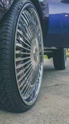 A close-up of a car wheel with a large, shiny rim featuring intricate designs. The tire has a deep tread pattern, and the background includes part of the car body and a hint of grass and pavement.