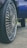 A close-up of a car wheel with a large, shiny rim featuring intricate designs. The tire has a deep tread pattern, and the background includes part of the car body and a hint of grass and pavement.