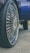 A close-up of a car wheel with a large, shiny rim featuring intricate designs. The tire has a deep tread pattern, and the background includes part of the car body and a hint of grass and pavement.