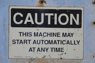 A rectangular caution sign with the word 'CAUTION' in large, bold letters at the top against a black background, followed by the text 'THIS MACHINE MAY START AUTOMATICALLY AT ANY TIME' in capital letters against a white background. The sign is mounted on a surface with signs of rust and wear.