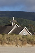 A small wooden chapel with a cross on its roof is surrounded by grass-covered sand dunes. The backdrop consists of dense green forest under a partly cloudy sky.