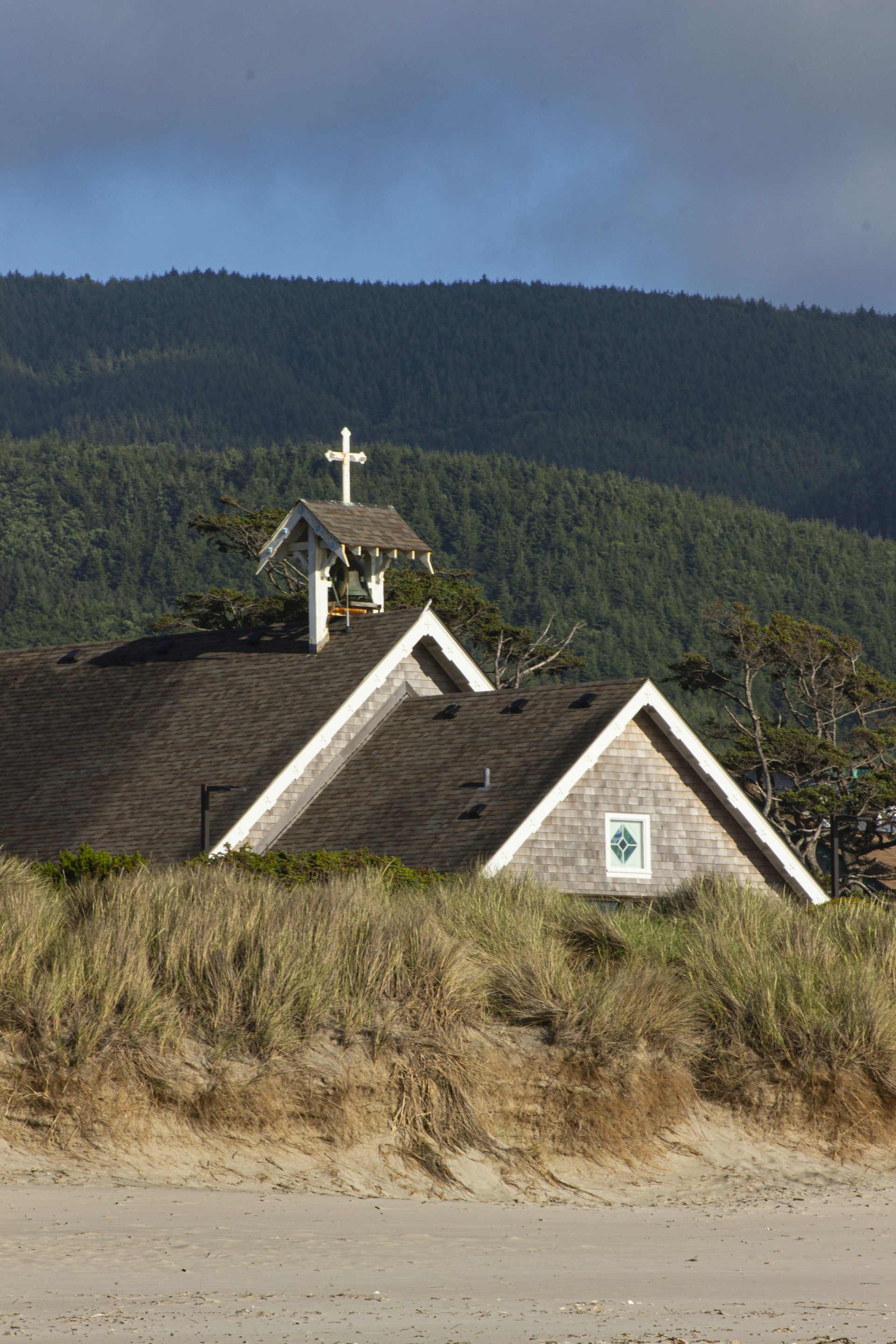 A small wooden chapel with a cross on its roof is surrounded by grass-covered sand dunes. The backdrop consists of dense green forest under a partly cloudy sky.