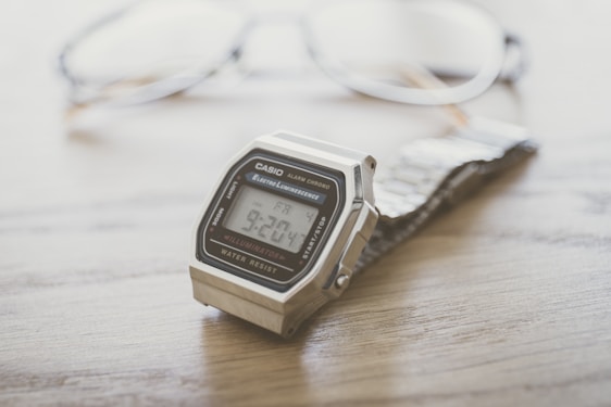 A Casio digital wristwatch with a metal band is placed on a wooden surface in focus, while a pair of eyeglasses is blurred in the background.