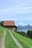 A rustic barn with a red-tiled roof sits atop a grassy hill overlooking a serene lake with distant mountains. A narrow dirt path leads up to the barn, flanked by lush green grass. The sky is clear with a few scattered clouds, adding to the tranquil, picturesque scenery.