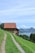 A rustic barn with a red-tiled roof sits atop a grassy hill overlooking a serene lake with distant mountains. A narrow dirt path leads up to the barn, flanked by lush green grass. The sky is clear with a few scattered clouds, adding to the tranquil, picturesque scenery.