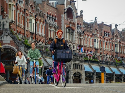 A group of people are riding bicycles along a city street lined with historic, ornate brick buildings. The architecture features arched windows and decorative stonework. The cyclists include both men and women, one of whom has a bike basket in front. The scene conveys a vibrant urban atmosphere.