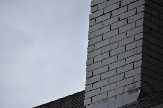 A close-up view of a brick chimney against an overcast sky. The chimney is constructed with uniform, rectangular bricks in a grayish color. The top part of a roof is visible, meeting the chimney at an angle.