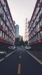 A narrow urban street is flanked by two rows of buildings, each with rows of windows and air conditioning units. Parked cars line the sides of the street. An electrical tower and power lines are visible in the background, against a partially cloudy sky.