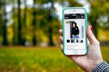 A hand holding a smartphone displaying an online shopping webpage. The screen shows a listing for a unisex sweatshirt with multiple images of a person modeling the garment. The background is a blurred scene of trees and green grass, suggesting an outdoor setting.