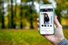 A hand holding a smartphone displaying an online shopping webpage. The screen shows a listing for a unisex sweatshirt with multiple images of a person modeling the garment. The background is a blurred scene of trees and green grass, suggesting an outdoor setting.