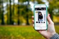 A hand holding a smartphone displaying an online shopping webpage. The screen shows a listing for a unisex sweatshirt with multiple images of a person modeling the garment. The background is a blurred scene of trees and green grass, suggesting an outdoor setting.