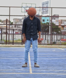 A person stands on an outdoor basketball court with a basketball positioned in front of their head, obscuring their face. They are wearing a black shirt, light blue jeans, and sandals. In the background, a basketball hoop and urban buildings are visible.
