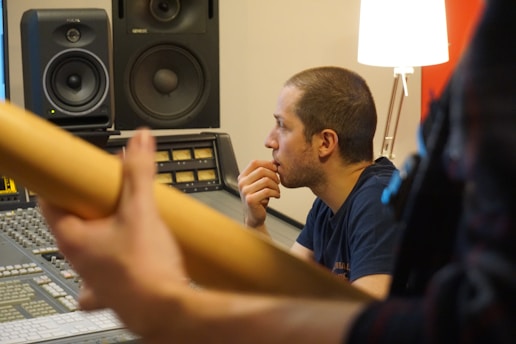 A man is seated at a mixing console in a recording studio, appearing focused and thoughtful while looking at a computer screen. A guitar is in the foreground with a large speaker and a lamp nearby.