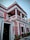 A two-story colonial-style building with pink and white exterior trim, featuring ornate architectural details and columns. It has a black door with the number 15 and decorative wall embellishments. Potted plants line the front of the building.