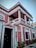 A two-story colonial-style building with pink and white exterior trim, featuring ornate architectural details and columns. It has a black door with the number 15 and decorative wall embellishments. Potted plants line the front of the building.