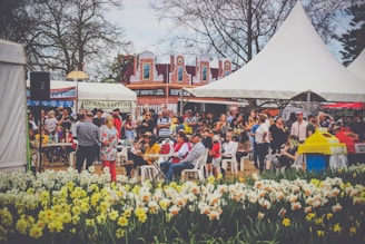 A lively outdoor market with people gathered around various stalls, including a henna tattoo booth and food stands. The setting is festive with colorful decorations and large tents. People are seated on white chairs, engaging in conversation and enjoying the sunny weather. In the foreground, a bed of vibrant yellow and white flowers can be seen, adding to the cheerful atmosphere.