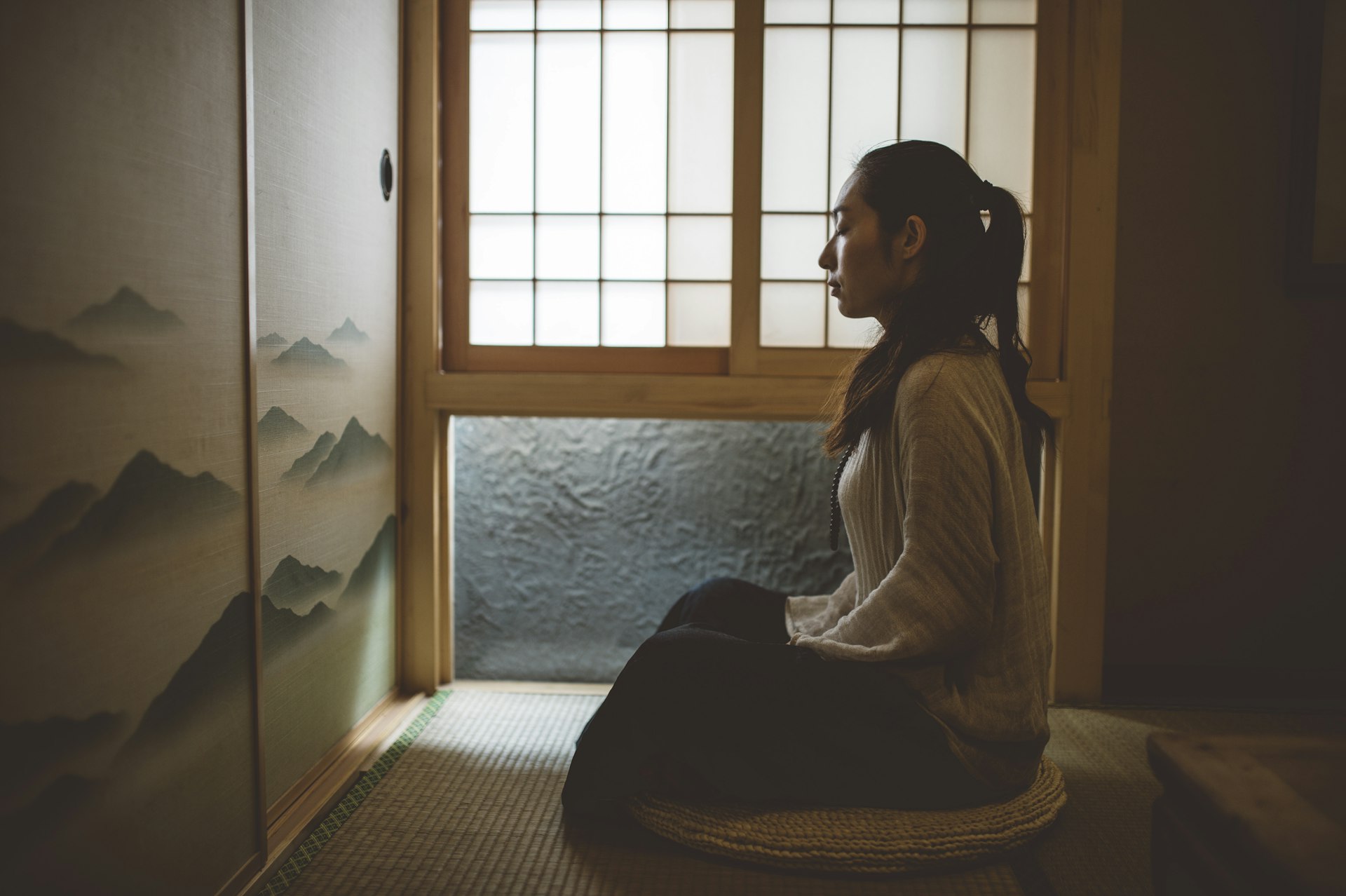 Woman kneeling on a round floor cushion beside a window in a quiet room