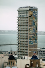 A tall, modern hotel building with numerous balconies and windows is prominently featured. The facade showcases a large, colorful mural with abstract and geometric designs. In the foreground, there are two traditional buildings with dome roofs, and the background reveals a calm sea with a dock.