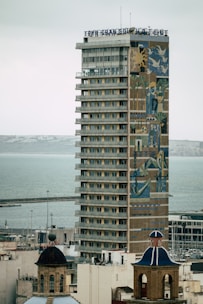 A tall, modern hotel building with numerous balconies and windows is prominently featured. The facade showcases a large, colorful mural with abstract and geometric designs. In the foreground, there are two traditional buildings with dome roofs, and the background reveals a calm sea with a dock.
