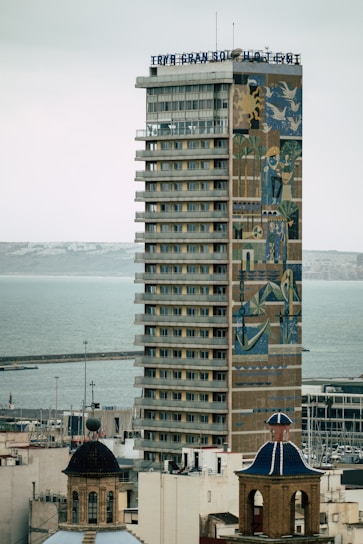 A tall, modern hotel building with numerous balconies and windows is prominently featured. The facade showcases a large, colorful mural with abstract and geometric designs. In the foreground, there are two traditional buildings with dome roofs, and the background reveals a calm sea with a dock.