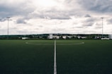 A clear, well-maintained soccer field with a central white line divides the green artificial turf. A backdrop of trees and buildings stretches across the horizon under a dramatic, cloudy sky.