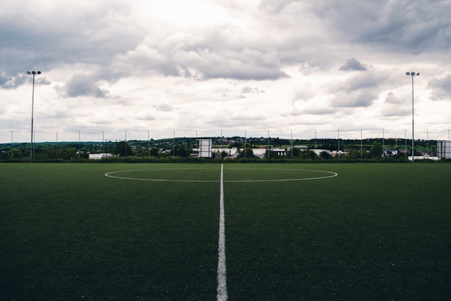 A clear, well-maintained soccer field with a central white line divides the green artificial turf. A backdrop of trees and buildings stretches across the horizon under a dramatic, cloudy sky.