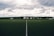 A clear, well-maintained soccer field with a central white line divides the green artificial turf. A backdrop of trees and buildings stretches across the horizon under a dramatic, cloudy sky.