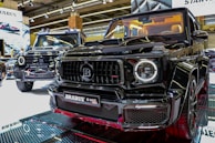 Two luxury black SUVs are displayed in a showroom under bright overhead lighting. The focus is on the front grill of one vehicle with a Brabus logo prominently featured. The interior of the closer SUV is illuminated, showcasing cream-colored seats.