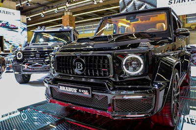 Two luxury black SUVs are displayed in a showroom under bright overhead lighting. The focus is on the front grill of one vehicle with a Brabus logo prominently featured. The interior of the closer SUV is illuminated, showcasing cream-colored seats.