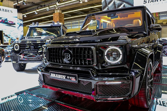 Two luxury black SUVs are displayed in a showroom under bright overhead lighting. The focus is on the front grill of one vehicle with a Brabus logo prominently featured. The interior of the closer SUV is illuminated, showcasing cream-colored seats.