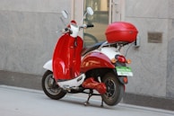 A red and white electric scooter is parked on a sidewalk next to a building. It has a top case for storage and a green license plate indicating it is an electric bike. The building wall is gray and has a modern, sleek texture.