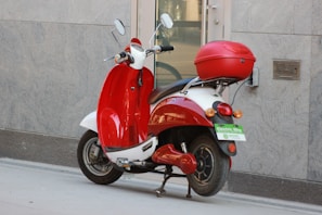 A red and white electric scooter is parked on a sidewalk next to a building. It has a top case for storage and a green license plate indicating it is an electric bike. The building wall is gray and has a modern, sleek texture.