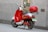 A red and white electric scooter is parked on a sidewalk next to a building. It has a top case for storage and a green license plate indicating it is an electric bike. The building wall is gray and has a modern, sleek texture.