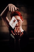A person with red hair and freckles focuses intensely on a playing card seemingly suspended in mid-air between their raised hands. The background is dark, adding to the mysterious atmosphere.