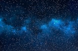 A vast expanse of deep space filled with countless stars against a backdrop of dark blue and black. Wisps of nebulae, glowing in soft shades of blue, seem to drift among the stars, creating a sense of depth and mystery.