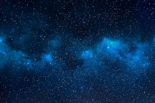 A vast expanse of deep space filled with countless stars against a backdrop of dark blue and black. Wisps of nebulae, glowing in soft shades of blue, seem to drift among the stars, creating a sense of depth and mystery.