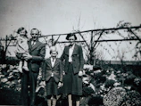 A vintage photograph features a family of four posing in a garden. The man on the left is holding a young child. A boy and a woman stand beside them. The background includes bushes and a wooden trellis with climbing plants.