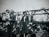 A vintage photograph features a family of four posing in a garden. The man on the left is holding a young child. A boy and a woman stand beside them. The background includes bushes and a wooden trellis with climbing plants.